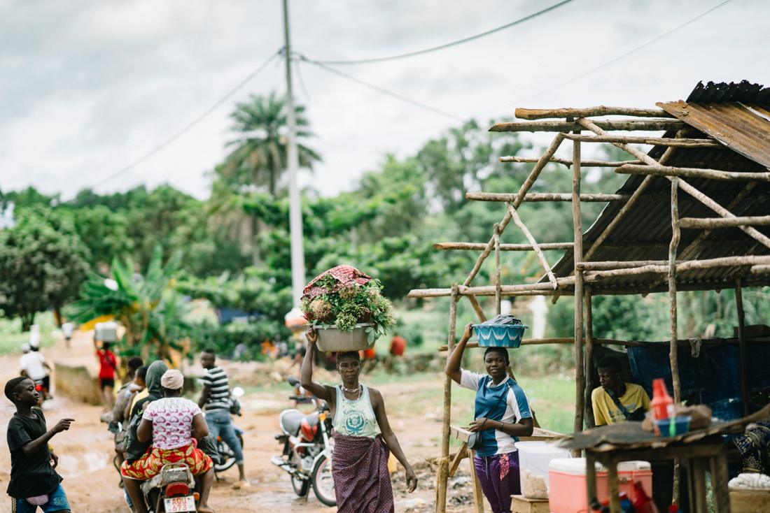 Women carrying produce on their heads along a road beside a market stall